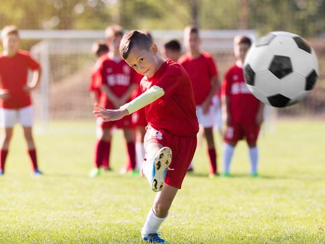 jonge jongen die een voetbal schopt terwijl zijn team op de achtergrond toekijkt