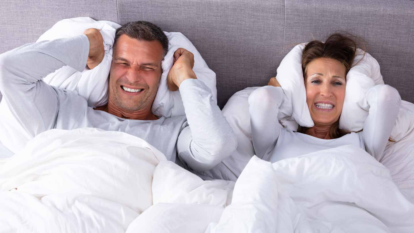 Marido y mujer en la cama con almohadas sobre la cabeza para bloquear a los vecinos ruidosos